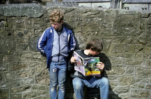 Warren and Luke rest by Gidleigh hostel wall; Luke engrossed in CRASH, the ZX Spectrum games magazine, with the church beyond.