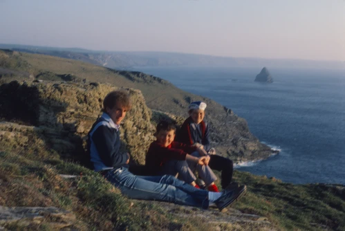 22 Apr 1987 · Michael, Mark and Jason Morris lounging outside Tintagel Youth Hostel on the grassy clifftop; Gull Rock beyond.