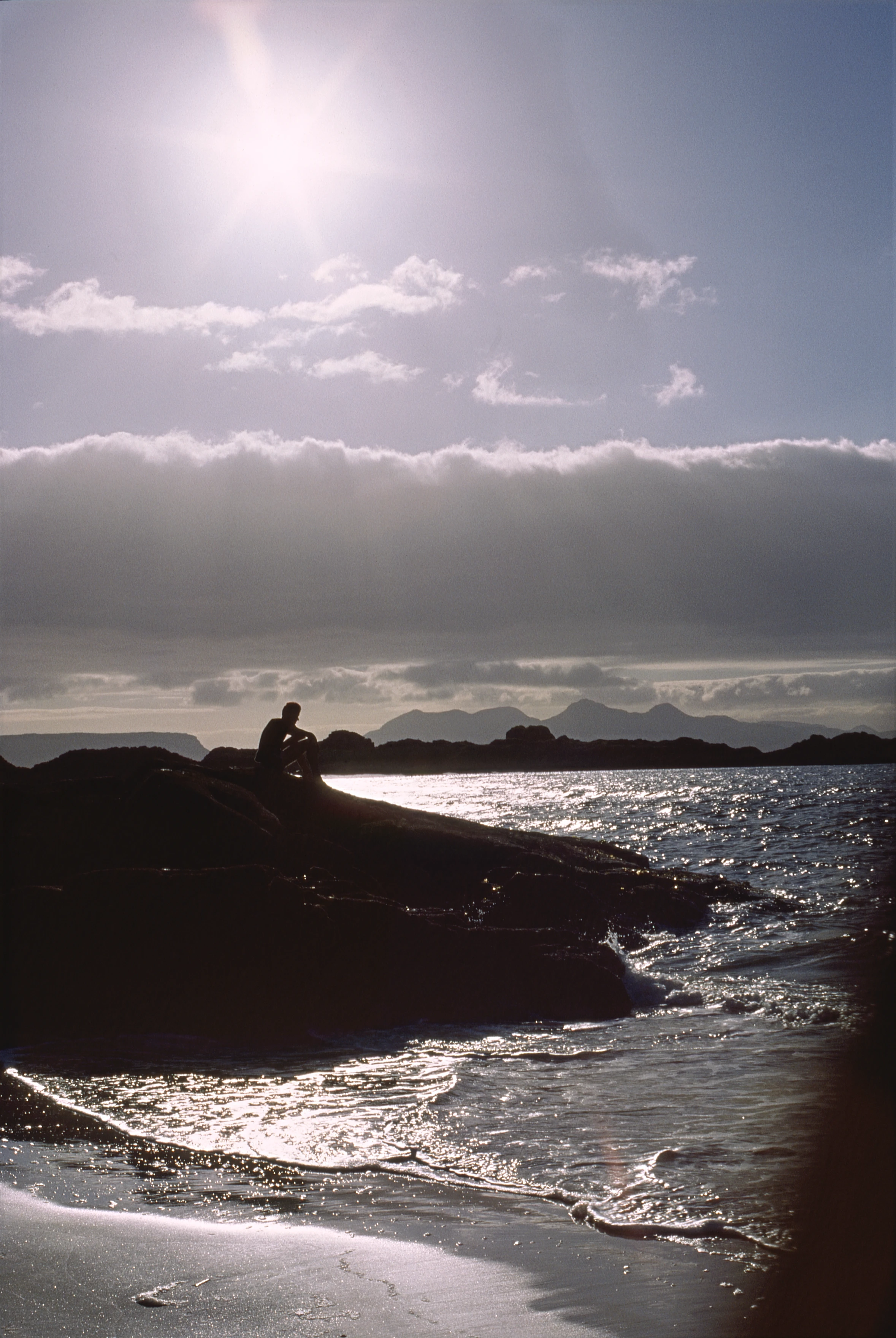 Michael Hall silhouetted on Camusdarach's south-west rocks, sun flaring on the tide. Part of our pre-supper beach walk.